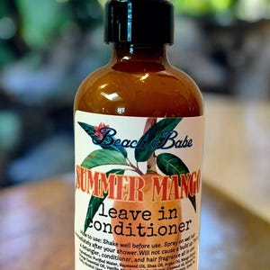 May include: A brown glass bottle with a black spray nozzle and a white label that reads "Beach Babe Summer Mango leave in conditioner". The label also includes instructions for use and a list of ingredients. The bottle is sitting on a wooden surface.