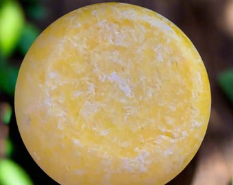May include: A round, yellow bar of shampoo with a white, grainy texture.