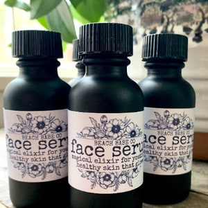 Puede incluir: Tres botellas de vidrio negro con tapas de rosca negras y etiquetas blancas. Las etiquetas tienen un dise&ntilde;o floral y el texto "BEACH BABE CO face serum magical elixir for youthful healthy skin that glows".