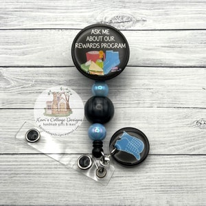 May include: A black and blue retractable badge reel with a black and white image of a house and the text "Kari's Cottage Designs handmade gifts & more Kariscottagedesigns@gmail.com". The top of the reel has a black circle with the text "ASK ME ABOUT OUR REWARDS PROGRAM" and a colorful image of a mug, yarn, socks, and a sandwich. The bottom of the reel has a black circle with a blue image of a pair of socks.