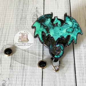 May include: A teal and black dragon-shaped retractable badge holder with glitter accents. The holder is attached to a clear plastic badge reel with a silver metal clip.