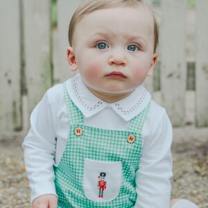 May include: A baby wearing a white long-sleeved shirt with a green gingham overall. The overall has a pocket with a nutcracker design and is buttoned with two brown buttons. The shirt has a white collar with red and green stitching.