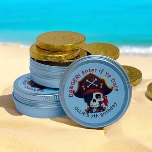 Puede incluir: Latas plateadas con un diseño de calavera de pirata y el texto "DANGER! Enter if Ye Dare Ollie's 7th Birthday" en la tapa. Las latas están llenas de monedas de chocolate doradas y están sobre una playa de arena.