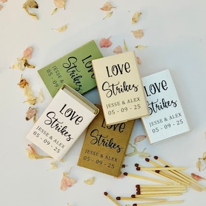Può includere: Una collezione di scatole di fiammiferi personalizzate in vari colori, tra cui bianco, beige e verde, con la scritta "Love Strikes" e una data. Fiammiferi sparsi e petali di fiori.
