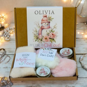 Peut inclure: Une boîte cadeau marron avec un couvercle blanc illustrant un bonhomme de neige et le texte "OLIVIA IT'S TIME TO Chill out!" La boîte contient une écharpe en fourrure blanche avec le texte "WARM your Mistle-Toes!" un pompon en fourrure rose, un petit pot de baume à lèvres avec une illustration de bonhomme de neige et le texte "Hot Cocoa Buds", et un petit pot de baume à lèvres avec une illustration de bonhomme de neige et le texte "From Kisses X".
