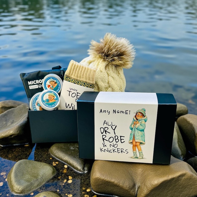 Pu&ograve; includere: Una confezione regalo con un'etichetta bianca che recita "Any Name! All Dry Robe & No Knickers" e un'illustrazione a fumetti. La scatola contiene un cappello a maglia a trecce color crema con un pompon, calzini e piccole scatole. &Egrave; visibile anche una borsa nera con la scritta "Micro Towel".