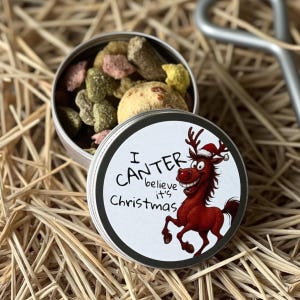 May include: A small, round tin filled with colorful treats, topped with a lid featuring a cartoon reindeer wearing a Santa hat. The lid reads "I can't believe it's Christmas."