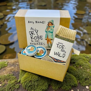 Könnte beinhalten: Eine beige Geschenkbox mit Socken, kleinen Dosen und einer Karte. Der Deckel der Box zeigt eine Zeichnung einer Person in einem Mantel und den Text "Any Name! All Dry Robe & No Knickers". Auf der Karte steht "you're TOE-tally WILD".