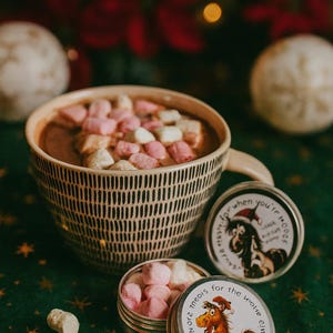 May include: A mug of hot chocolate topped with pink and white marshmallows, next to two small tins with cartoon horse and dog illustrations. The mug has a black and white pattern and is on a green surface.