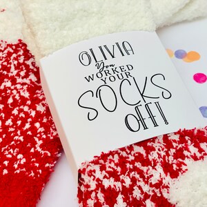 以下が含まれることがあります： 赤と白のふわふわの靴下で、白いタグには「Olivia you worked your socks off!」と書かれています。