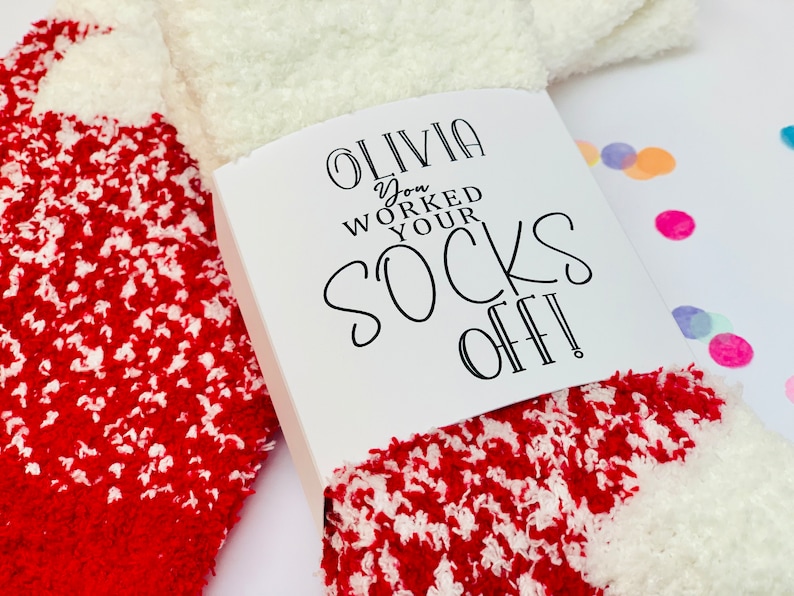 以下が含まれることがあります： 赤と白のふわふわの靴下で、白いタグには「Olivia you worked your socks off!」と書かれています。