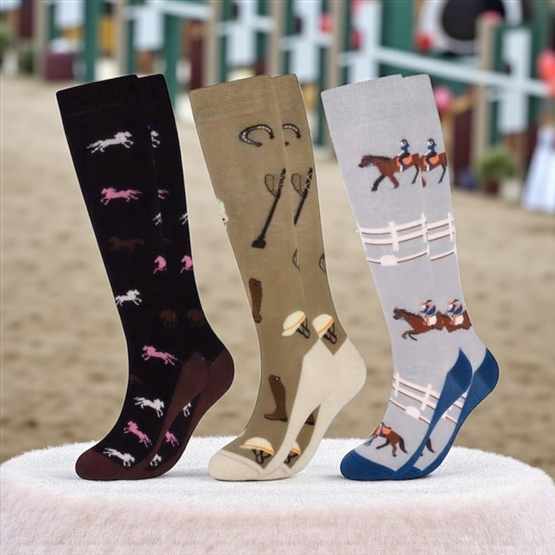 May include: Three pairs of knee-high socks with equestrian-themed designs. One pair is black with pink and white horse silhouettes, another is tan with riding gear, and the last is gray with riders and fences.