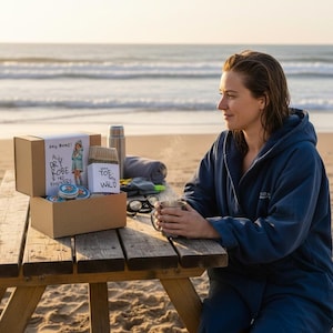 Pu&ograve; includere: Una persona con un accappatoio blu &egrave; seduta a un tavolo da picnic di legno su una spiaggia, con in mano una tazza. Una scatola di cartone con testo e illustrazioni &egrave; sul tavolo, insieme a un thermos, un asciugamano e altri oggetti. La scena &egrave; ambientata sulla spiaggia con il mare sullo sfondo.