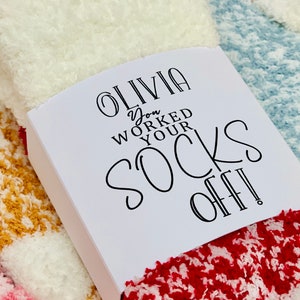 以下が含まれることがあります： 白いギフトタグに黒い文字で「Olivia You Worked Your Socks Off!」と書かれています。タグは、赤、白、青のふわふわした靴下の山の上に置かれています。