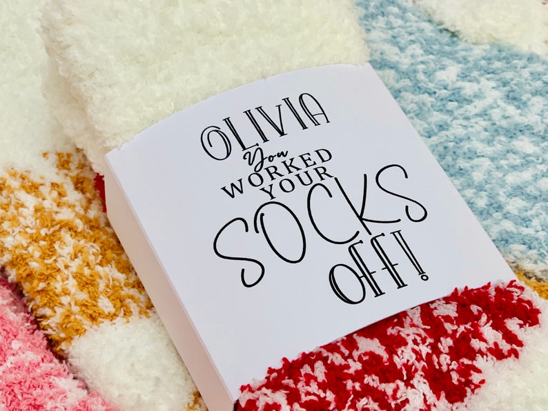 以下が含まれることがあります： 白いギフトタグに黒い文字で「Olivia You Worked Your Socks Off!」と書かれています。タグは、赤、白、青のふわふわした靴下の山の上に置かれています。
