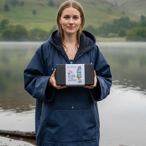 Pu&ograve; includere: Un accappatoio con cappuccio blu navy con fodera in pile, tenuto da una persona vicino a un lago. L'accappatoio ha una tasca frontale e un cappuccio con coulisse. Una scatola nera con la scritta "A Dry Robe & No Worries" &egrave; tenuta davanti all'accappatoio.