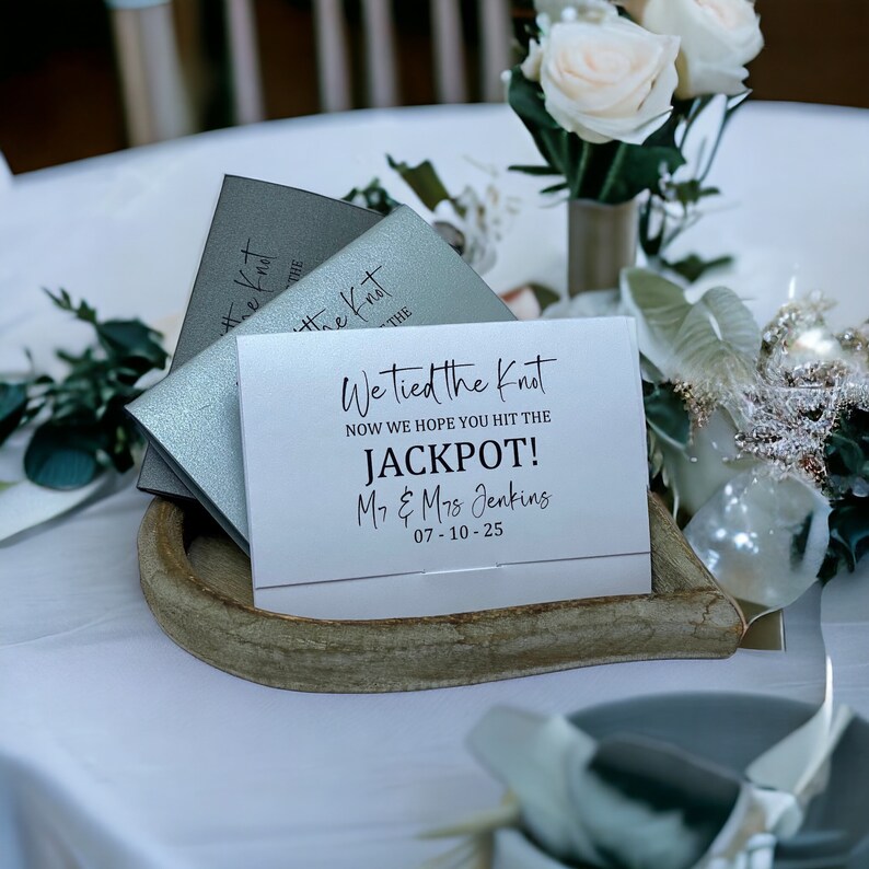 Puede incluir: Una tarjeta blanca con texto negro que dice "We tied the knot now we hope you hit the JACKPOT! Mr. & Mrs. Jenkins 07-10-25". La tarjeta est&aacute; sobre una bandeja de madera con dos cajas plateadas detr&aacute;s.