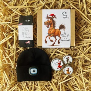 May include: A gift set with a Christmas theme, featuring a black beanie with a light, a pair of black socks with horse graphics, and a greeting card with a cartoon horse wearing a Santa hat. The set also includes three small tins.