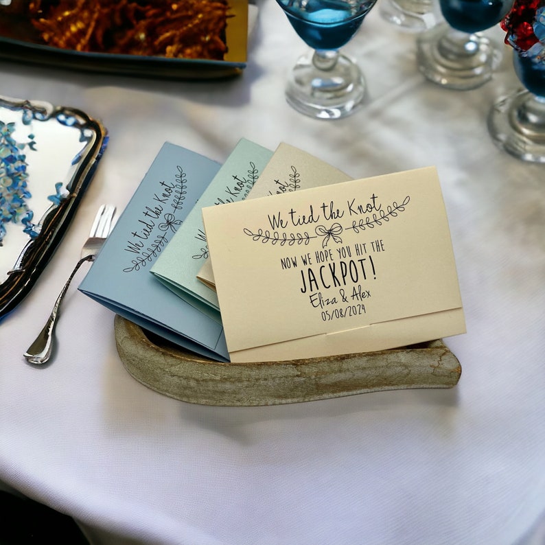 Puede incluir: Una tarjeta de color amarillo claro con el texto "We tied the knot" y "Now we hope you hit the jackpot!" con un lazo decorativo y un dise&ntilde;o floral. La tarjeta tambi&eacute;n incluye los nombres "Eliza & Alex" y la fecha "05/08/2024".