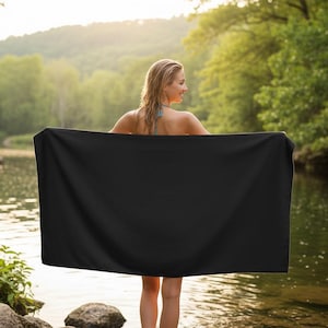 Pu&ograve; includere: Una persona in piedi in un fiume, che tiene un grande asciugamano rettangolare nero. Lo sfondo presenta una lussureggiante foresta verde e uno specchio d'acqua, con la luce del sole che filtra attraverso gli alberi.