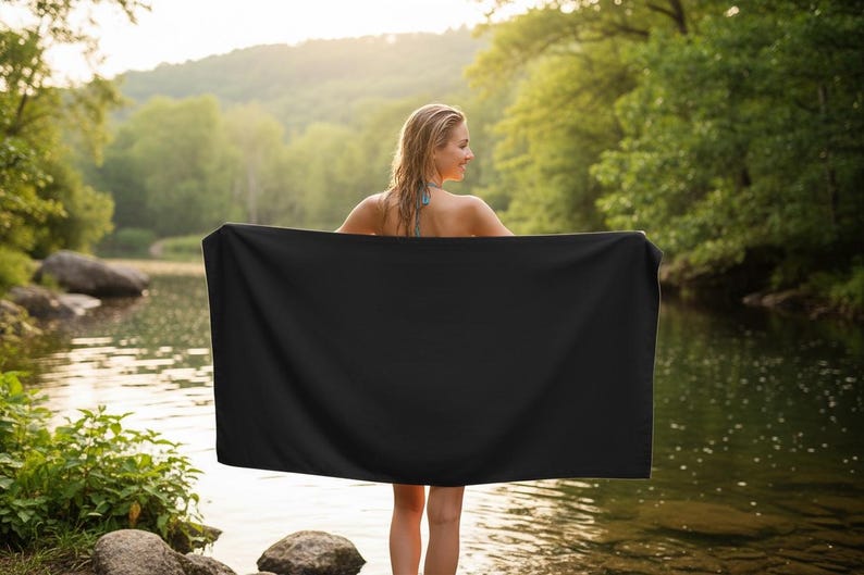 Pu&ograve; includere: Una persona in piedi in un fiume, che tiene un grande asciugamano rettangolare nero. Lo sfondo presenta una lussureggiante foresta verde e uno specchio d'acqua, con la luce del sole che filtra attraverso gli alberi.