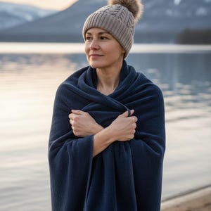Pu&ograve; includere: Una donna indossa un cappello di lana grigio con un pompon marrone e un asciugamano blu navy. La donna &egrave; in piedi vicino a un lago con montagne sullo sfondo. L'asciugamano &egrave; avvolto attorno alle sue spalle.
