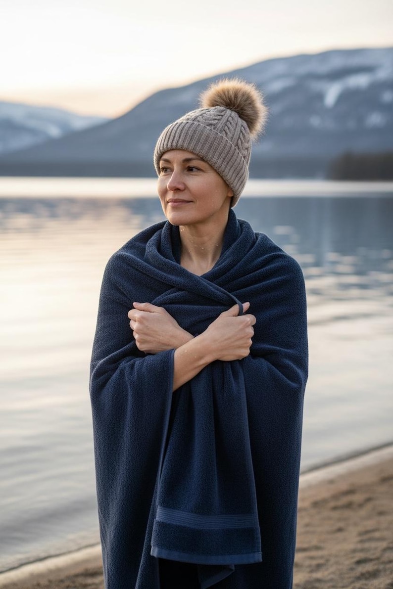 Pu&ograve; includere: Una donna indossa un cappello di lana grigio con un pompon marrone e un asciugamano blu navy. La donna &egrave; in piedi vicino a un lago con montagne sullo sfondo. L'asciugamano &egrave; avvolto attorno alle sue spalle.