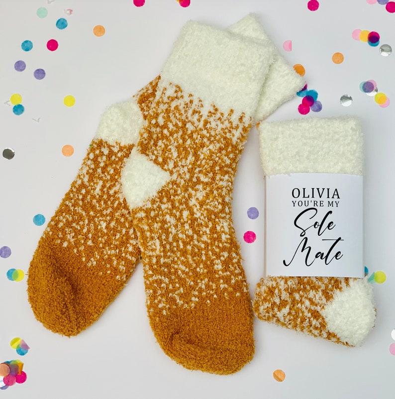 以下が含まれることがあります： 白とマスタードイエローのふわふわしたソックスのペアで、白いタグに「Olivia you're my Sole Mate」の文字が書かれています。