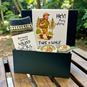 May include: A black gift box containing hiking-themed items. Includes a pair of patterned socks, a card with a hiker illustration and the text "Hey! Any NAME", and two round tins. The card reads "Take a walk" and "Because you've walked your socks".