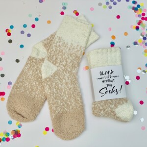 以下が含まれることがあります： ベージュと白のふわふわしたソックスのペア。タグには「Olivia Life Without You Socks!」と書かれています。