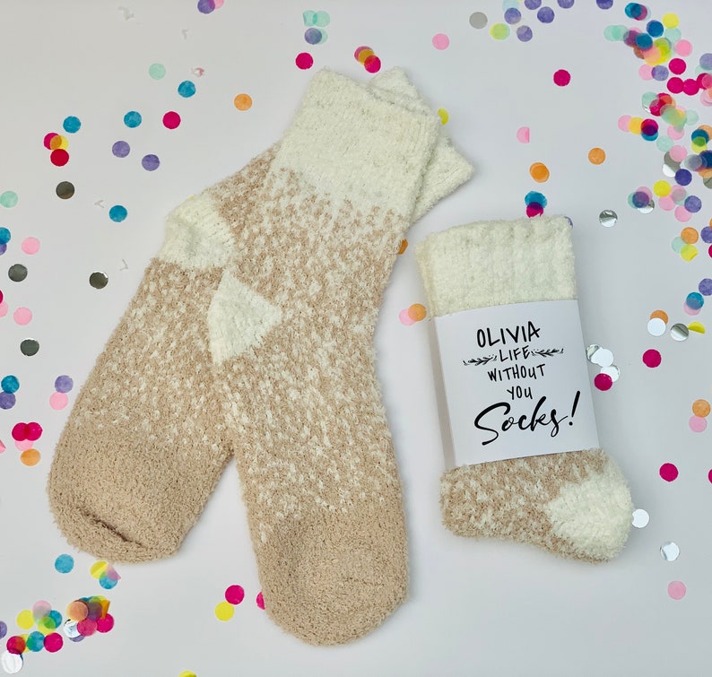 以下が含まれることがあります： ベージュと白のふわふわしたソックスのペア。タグには「Olivia Life Without You Socks!」と書かれています。
