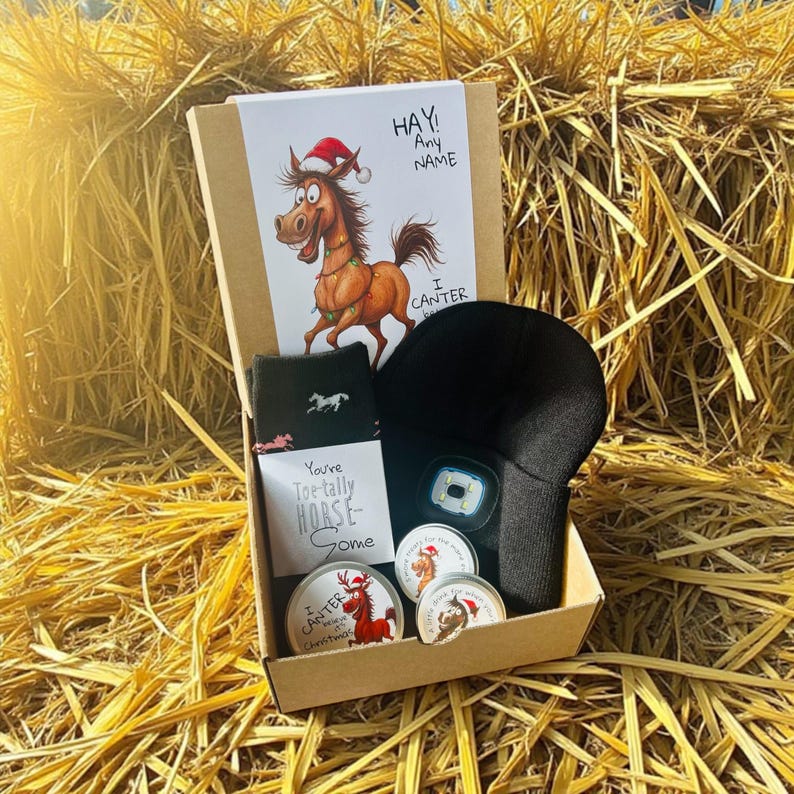 May include: A gift box with horse-themed items. Includes a card with a cartoon horse wearing a Santa hat, socks with a horse silhouette, a black headlamp, and round tins with horse illustrations. The box is on a hay bale.