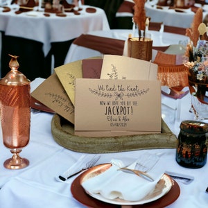 Puede incluir: Una tarjeta marr&oacute;n con el texto "We tied the knot. Now we hope you hit the jackpot! Eliza & Alex 05/08/2029" sobre una mesa con un jarr&oacute;n de cobre y un frasco de vidrio.