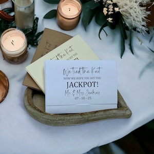 Puede incluir: Una tarjeta blanca con texto negro que dice "We tied the knot. Now we hope you hit the JACKPOT! Mr. & Mrs. Jenkins 07-10-25". La tarjeta est&aacute; sobre una bandeja de madera en forma de coraz&oacute;n.
