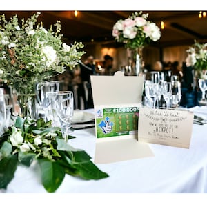 Puede incluir: Una mesa de recepci&oacute;n de boda con un mantel blanco, centros de mesa florales verdes y una tarjeta que dice "Nos hemos casado, ahora esperamos que te toque el premio gordo!" con un boleto de loter&iacute;a dentro de un sobre.