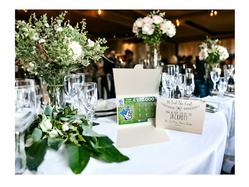 Puede incluir: Una mesa de recepci&oacute;n de boda con un mantel blanco, centros de mesa florales verdes y una tarjeta que dice "Nos hemos casado, ahora esperamos que te toque el premio gordo!" con un boleto de loter&iacute;a dentro de un sobre.