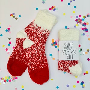 以下が含まれることがあります： 「Olivia You Worked Your Socks Off!」と書かれた白いタグが付いた、赤と白のふわふわしたソックスのペア