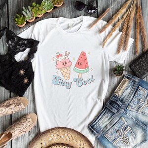 Puede incluir: Camiseta blanca con un gr&aacute;fico de un cono de helado y una paleta de sand&iacute;a con el texto "Stay Cool".