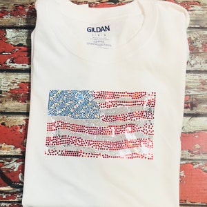 May include: White t-shirt with a red, white, and blue rhinestone American flag design. The shirt is a Gildan brand.