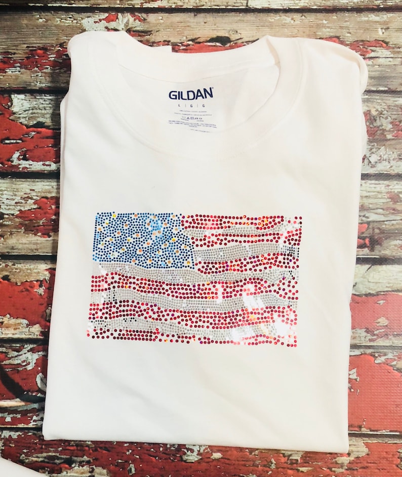 May include: White t-shirt with a red, white, and blue rhinestone American flag design. The shirt is a Gildan brand.