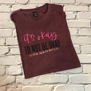 May include: Maroon t-shirt with the text "it's okay TO NOT BE OKAY MENTAL HEALTH MATTERS" in pink and black lettering. The shirt is folded and placed on a white brick wall.