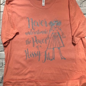 Puede incluir: Una camiseta de color melocot&oacute;n con un estampado gr&aacute;fico gris de una mujer con una expresi&oacute;n de sorpresa y el texto "Nunca subestimes el poder de un berrinche."