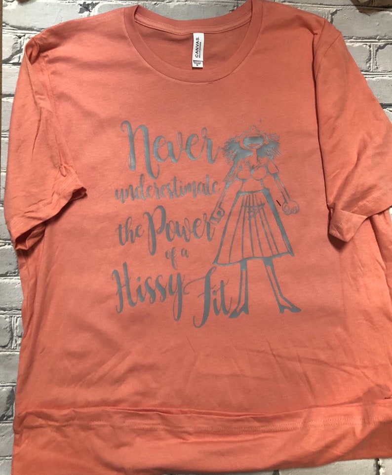 Puede incluir: Una camiseta de color melocot&oacute;n con un estampado gr&aacute;fico gris de una mujer con una expresi&oacute;n de sorpresa y el texto "Nunca subestimes el poder de un berrinche."