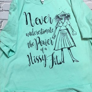 Puede incluir: Una camiseta verde claro con un estampado gr&aacute;fico negro de una mujer con una expresi&oacute;n de sorpresa. El texto en la camiseta dice "Never underestimate the power of a hissy fit."