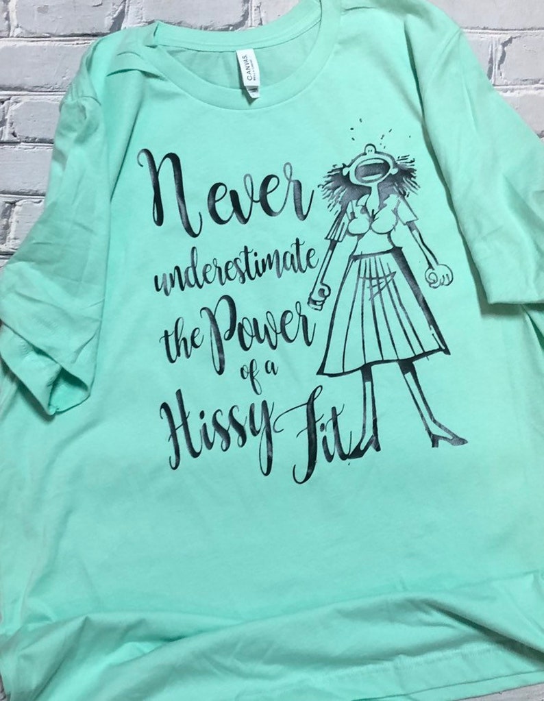 Puede incluir: Una camiseta verde claro con un estampado gr&aacute;fico negro de una mujer con una expresi&oacute;n de sorpresa. El texto en la camiseta dice "Never underestimate the power of a hissy fit."