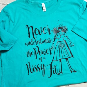 Puede incluir: Una camiseta turquesa con un estampado gr&aacute;fico negro de una mujer con los brazos en alto y el texto "Never underestimate the power of a hissy fit".