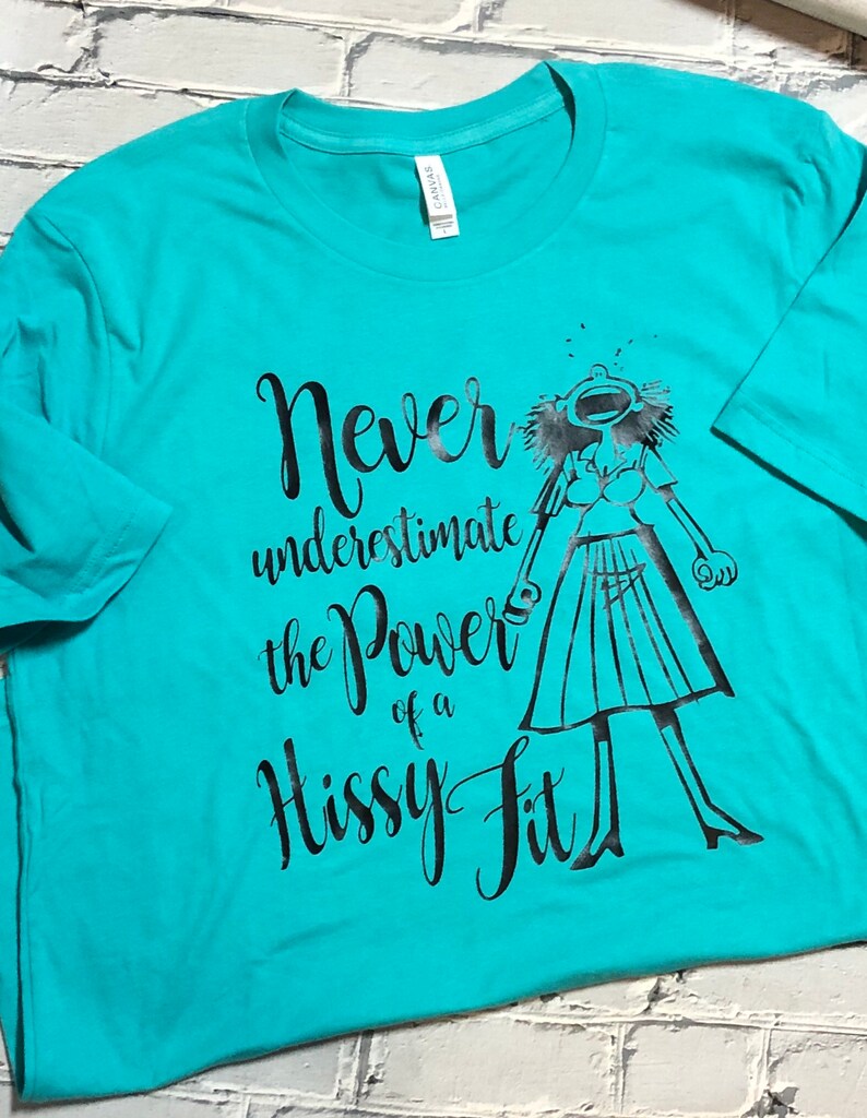 Puede incluir: Una camiseta turquesa con un estampado gr&aacute;fico negro de una mujer con los brazos en alto y el texto "Never underestimate the power of a hissy fit".
