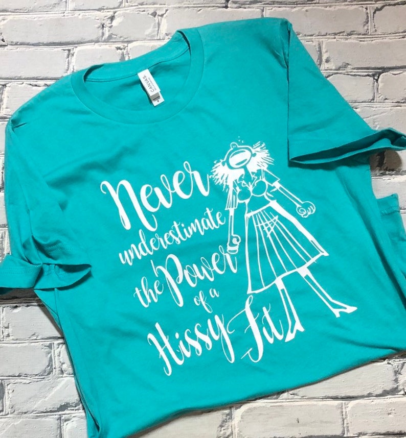Puede incluir: Una camiseta azul turquesa con un dise&ntilde;o gr&aacute;fico blanco. El gr&aacute;fico presenta a una mujer de dibujos animados con una expresi&oacute;n de sorpresa y el texto "Nunca subestimes el poder de un berrinche."