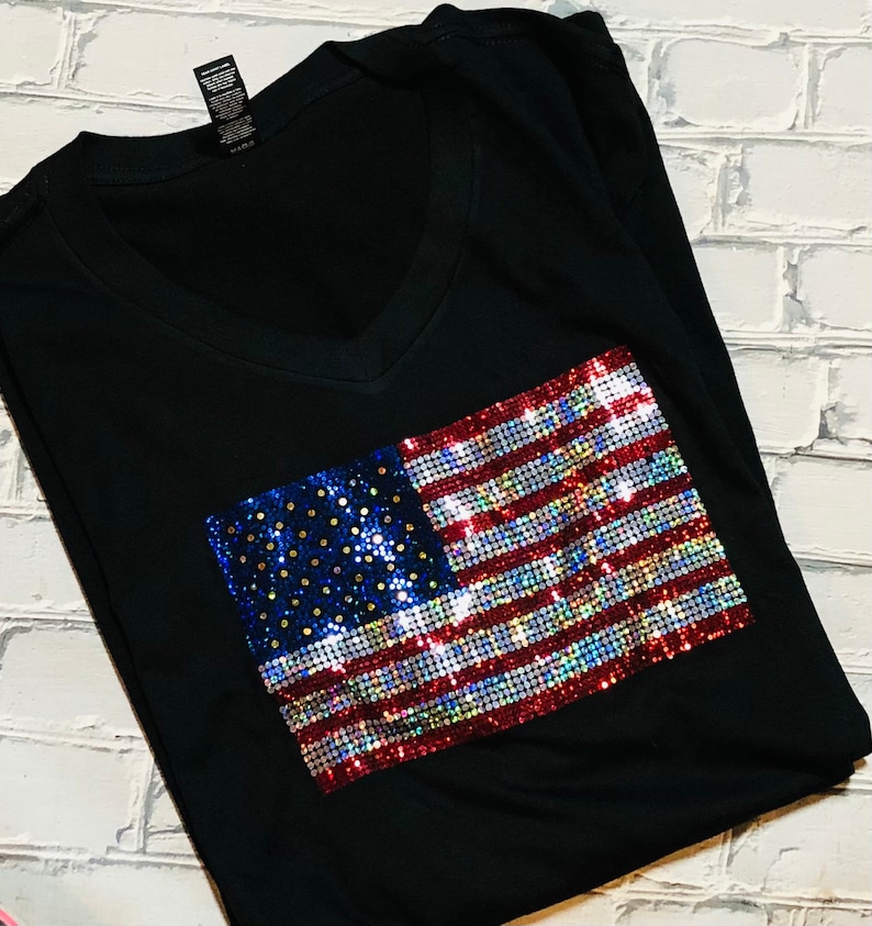 May include: A black v-neck t-shirt with a sequined American flag design. The flag is red, white, and blue with iridescent sequins.