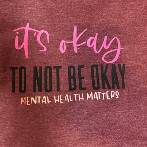 May include: A maroon t-shirt with the words "it's okay" in pink script. Below, in black block letters, it reads "TO NOT BE OKAY". The phrase "MENTAL HEALTH MATTERS" is printed in white beneath the black text.